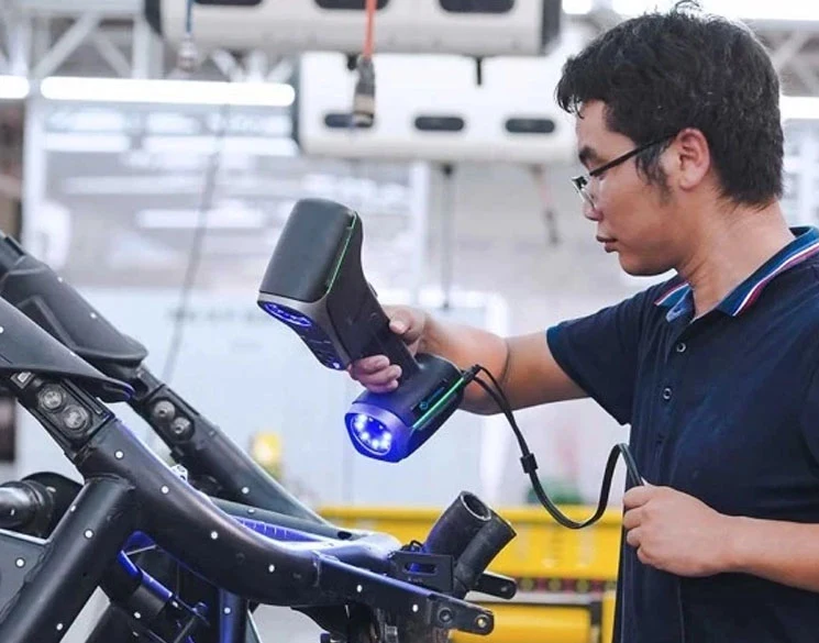 A technician in a workshop using a handheld Scanology 3D laser scanner to capture high-density surface data from a complex tubular metal chassis. The scanner emits a blue light as it moves over the dark industrial framework to create a precise digital model.